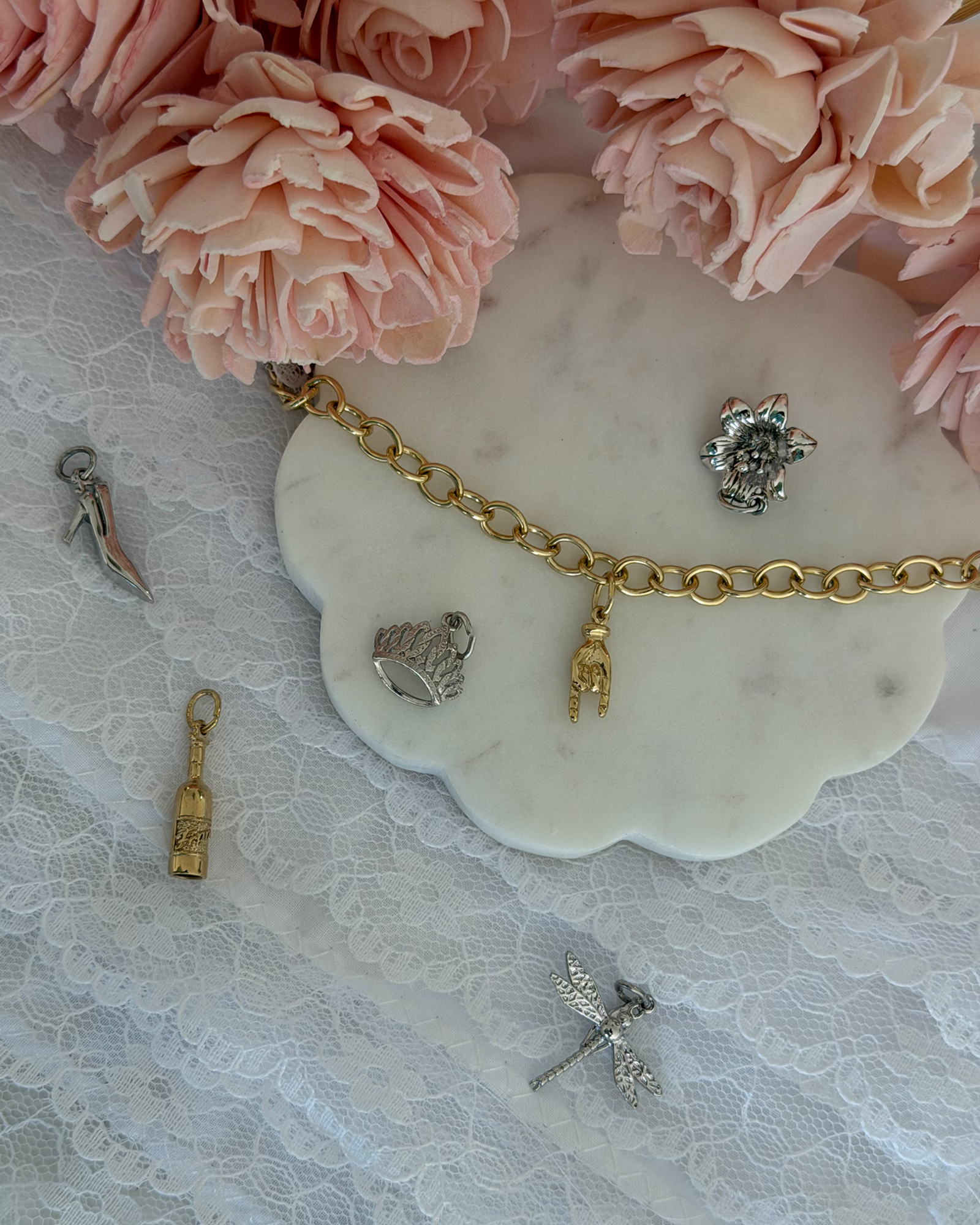 Gold charm bracelet with a hand charm on a marble tray, surrounded by high heel, crown, flower, bottle and dragonfly charms, pink flowers and lace