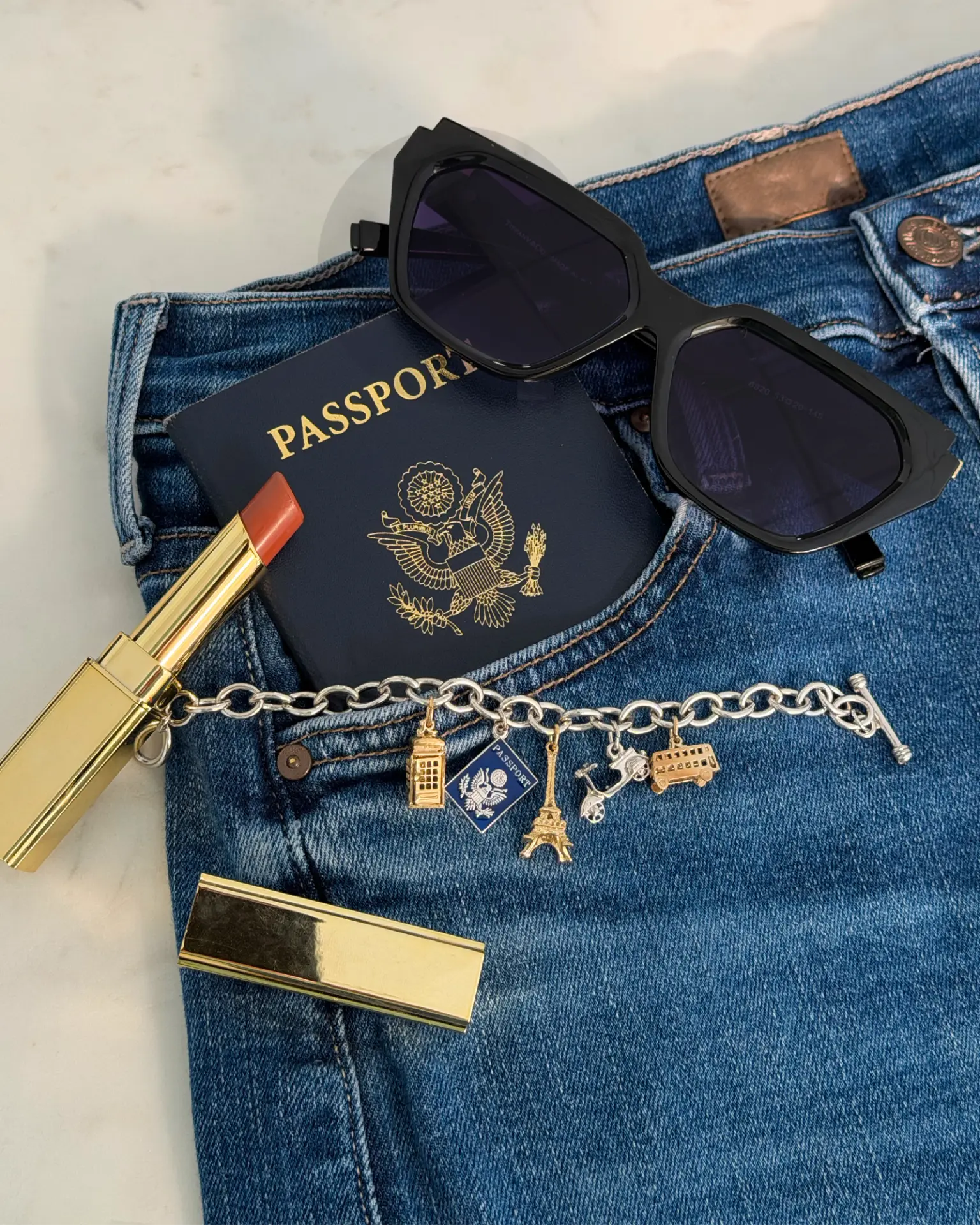 Silver charm bracelet with phone booth, passport, Eiffel Tower, scooter and double-decker bus charms on jeans beside a passport, sunglasses and lipstick