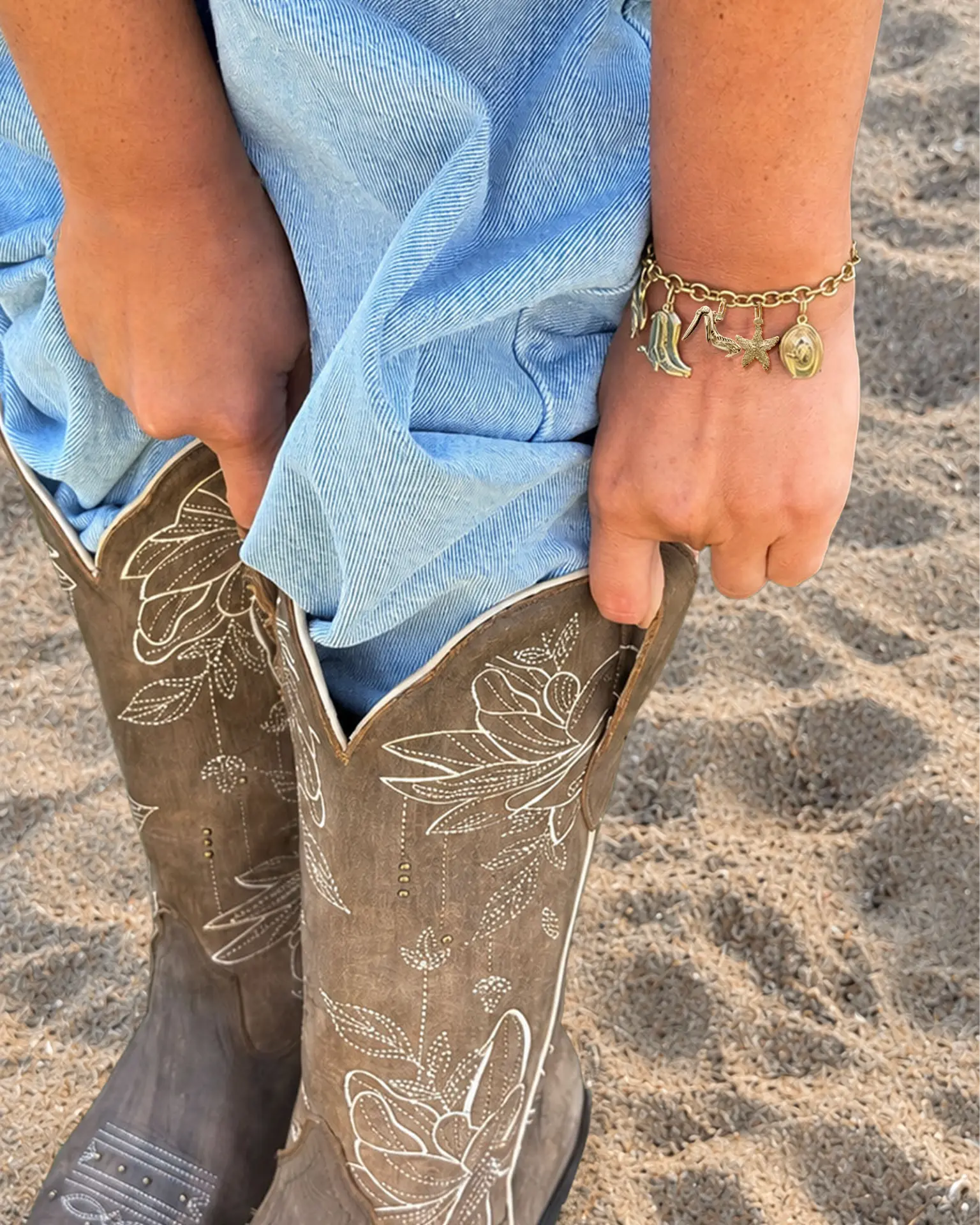 A person sitting on sand wearing denim shorts and floral-stitched cowboy boots, with a gold charm bracelet on the wrist