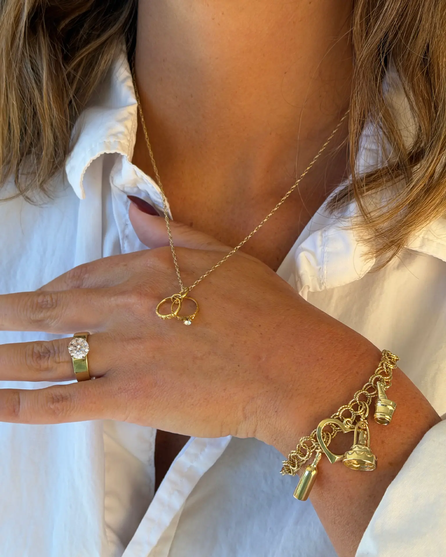 Woman in a white shirt wearing a linked wedding rings pendant and a gold charm bracelet with heart, wedding cake and champagne charms