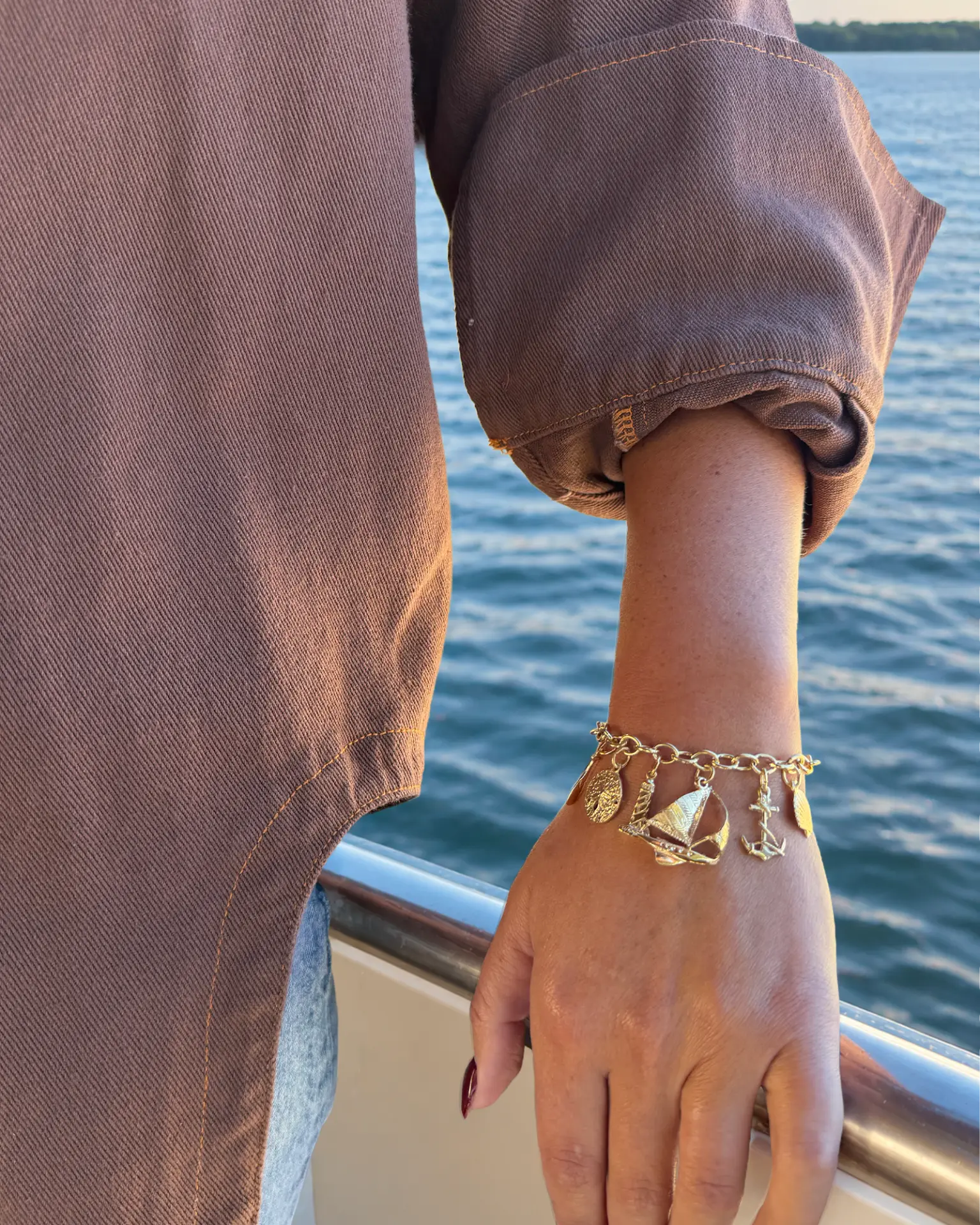 Wrist resting on a boat rail over the water, wearing a gold charm bracelet with sailboat, anchor, lighthouse and sand dollar charms