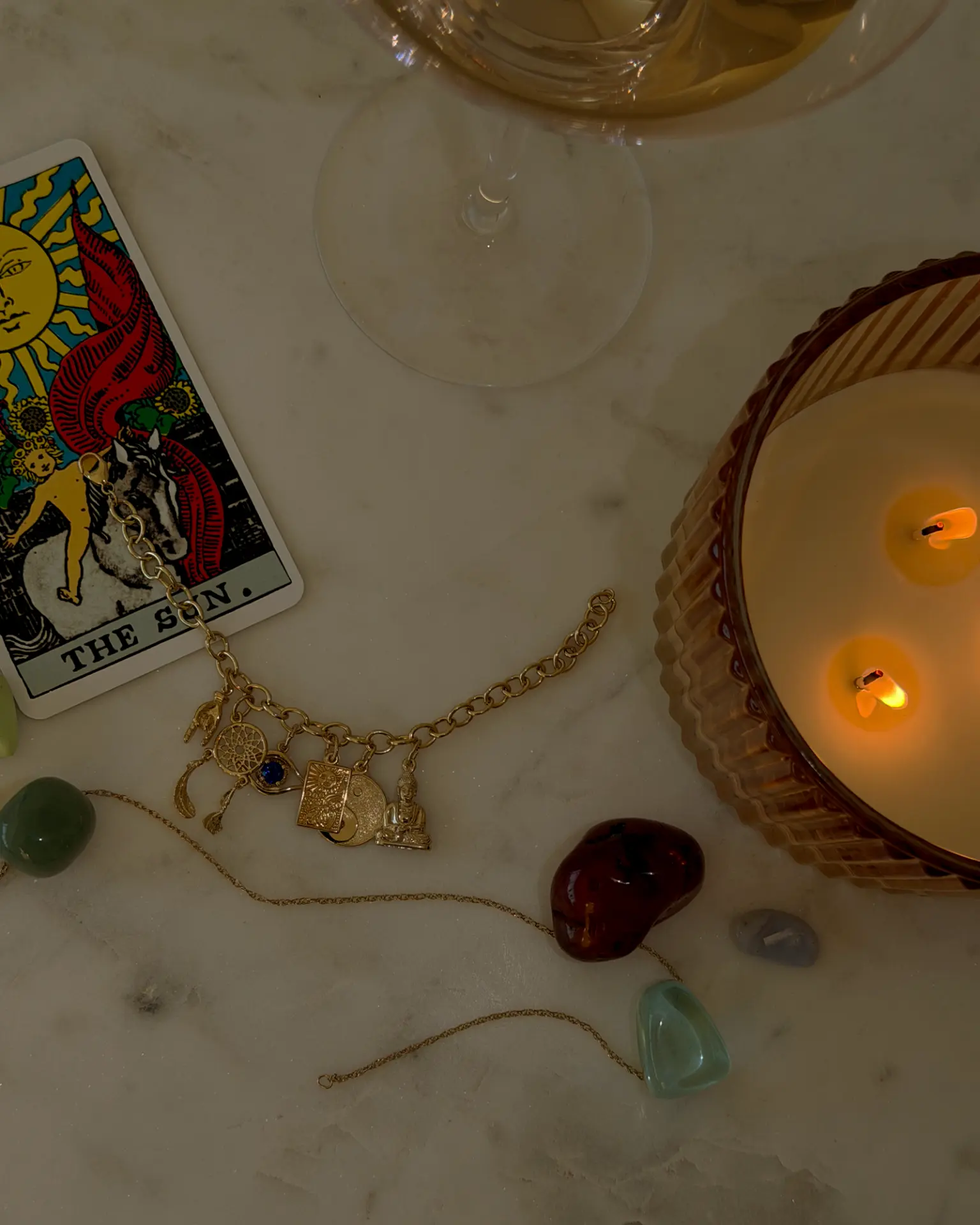 Gold charm bracelet with dreamcatcher, evil eye, sun tablet, yin yang and Buddha charms beside a tarot card, crystals and a lit candle