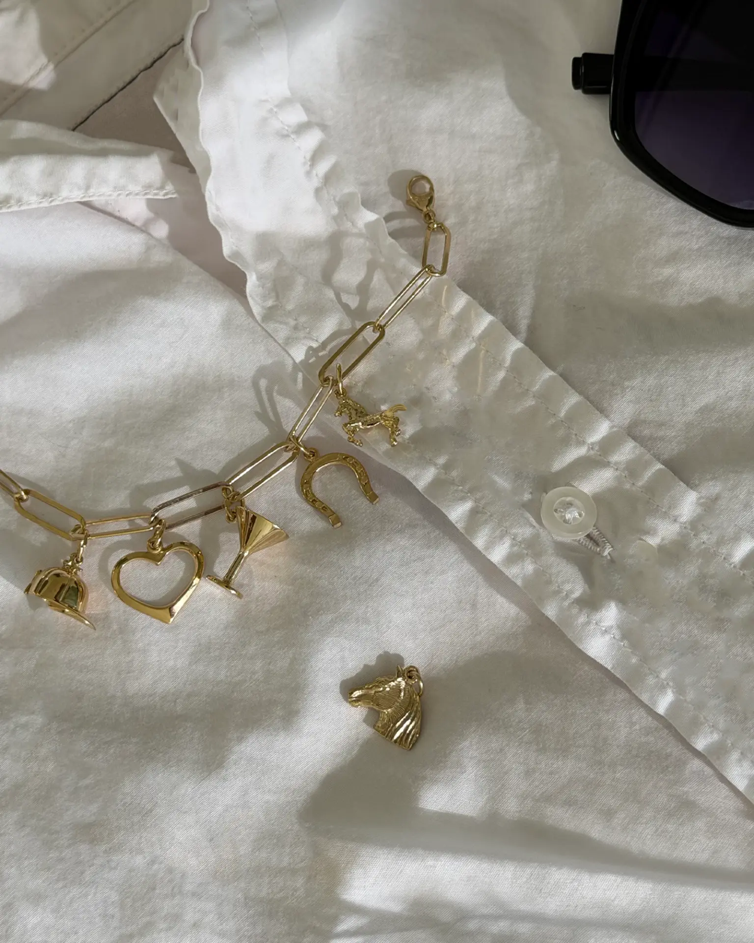Gold paperclip charm bracelet with riding cap, heart, martini glass, horseshoe and horse charms on a white shirt, a horse head charm beside it